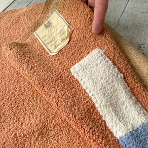 Può includere: Primo piano di una mano che tiene un pezzo di stoffa con un'etichetta che recita "Patron 1130, No. 215016, Largeur 1/2,  Mitaine 3/4,  Baller,  Deux Joues". Il tessuto è un mix di arancione, bianco e blu.