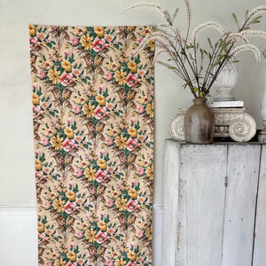 May include: A vintage floral fabric with a beige background and a repeating pattern of pink, yellow, and green flowers. The fabric is displayed against a white wall and a white cabinet.