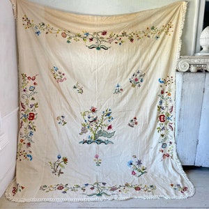 May include: A large, off-white linen tablecloth with a delicate floral embroidery design. The tablecloth features a central motif of a large floral arrangement with smaller floral motifs scattered throughout. The tablecloth has a white crocheted border.