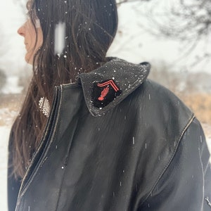 Puede incluir: Una chaqueta de cuero negra con un parche rojo y negro en el hombro. El parche presenta un pájaro estilizado con las alas extendidas.