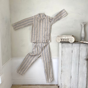 May include: A striped pajama set in beige, blue, and white. The top has a button-up front and long sleeves. The pants have a straight leg and are hanging on a white wall.