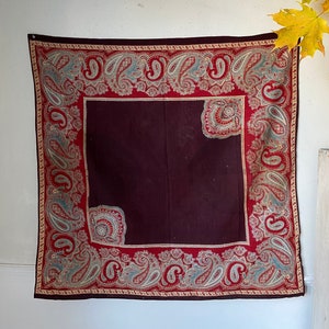 Puede incluir: Una bufanda cuadrada de color burdeos con estampado paisley y borde rojo y crema. La bufanda está colgada en una pared blanca.