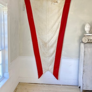 Può includere: Una bandiera bianca con due strisce rosse sui lati. La bandiera è appesa a un muro e ha un aspetto usurato e vintage.