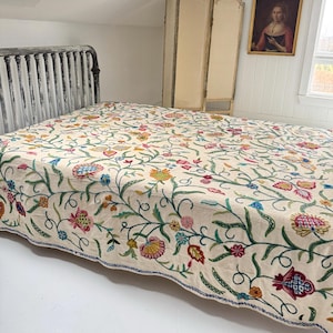 May include: A cream-colored bedspread with intricate floral embroidery in shades of pink, yellow, blue, and green. The bedspread is on a bed with a dark metal frame. A decorative screen and a framed portrait are in the background.
