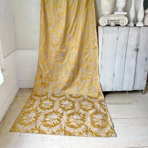 May include: A pair of gold and white damask curtains with a floral pattern. The curtains are hanging from a white wall and are draped on the floor.