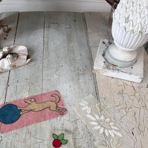 May include: A vintage fabric applique featuring a dog playing with a blue ball on a pink background. There is also a red berry with green leaves and a white floral lace trim on a distressed wooden surface.
