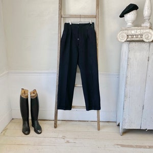 May include: Black high-waisted pants hanging on a wooden ladder. The pants have a single button closure and pleats at the front.
