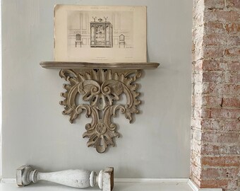 May include: A white painted shelf with ornate scrollwork details. The shelf is mounted to a white wall and has a framed print on top of it. The print is in black and white and shows a drawing of a room with furniture.