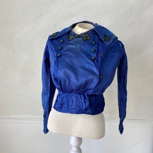 May include: A vintage cobalt blue jacket with a double-breasted design. The jacket features a wide collar, long sleeves, and decorative buttons. The collar and upper chest are adorned with embroidered embellishments. The jacket is displayed on a mannequin.