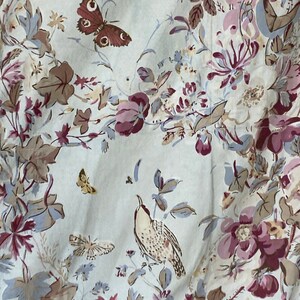 Puede incluir: Una tela blanca con un patr&oacute;n floral con flores rosas, marrones y azules, hojas y mariposas. Un peque&ntilde;o p&aacute;jaro est&aacute; posado en una rama.