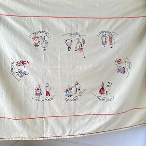 May include: A white tablecloth with red embroidered borders and colorful embroidered figures representing different regions of France. The text "Ile de France", "Bourgogne", "Franche Comte", "Alsace", "Dauphine", "Champagne", and "Normandie" is embroidered on the tablecloth.