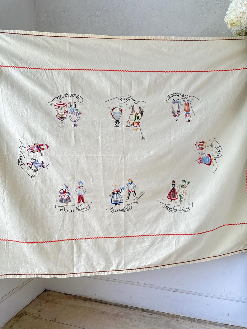 May include: A white tablecloth with red embroidered borders and colorful embroidered figures representing different regions of France. The text "Ile de France", "Bourgogne", "Franche Comte", "Alsace", "Dauphine", "Champagne", and "Normandie" is embroidered on the tablecloth.
