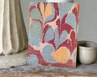 Può includere: Un piccolo libro rettangolare con una copertina marmorizzata che presenta motivi organici rossi, blu e gialli. È appoggiato su una superficie di legno invecchiato, con un piccolo vaso di ceramica e oggetti decorativi sullo sfondo.