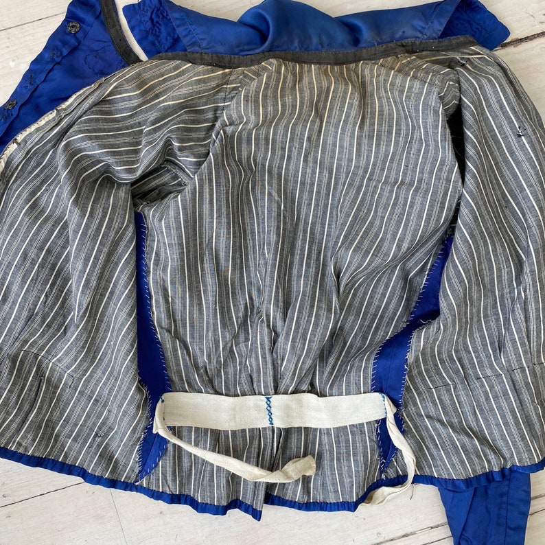 May include: A vintage blue jacket with a gray and white striped lining. The jacket has a white belt and blue stitching. The inside lining is a gray and white striped pattern. The jacket is open, showing the interior construction.