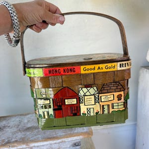 Könnte beinhalten: Ein Vintage-Picknickkorb aus Weide mit einem Holzgriff. Der Korb ist mit einer Szene von Häusern und dem Text "Good As Gold" und "Hong Kong" bemalt.