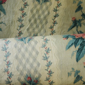 K&ouml;nnte beinhalten: Nahaufnahme eines cremefarbenen Stoffes mit einem sich wiederholenden Muster aus blauen und rosa Blumenmotiven. Der Stoff ist aufgerollt und zeigt das Muster sowohl auf der Ober- als auch auf der Unterseite der Rolle.