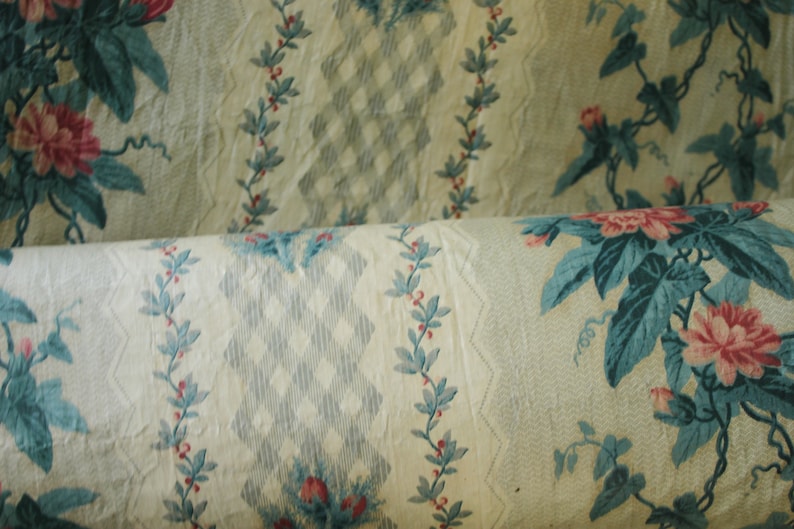K&ouml;nnte beinhalten: Nahaufnahme eines cremefarbenen Stoffes mit einem sich wiederholenden Muster aus blauen und rosa Blumenmotiven. Der Stoff ist aufgerollt und zeigt das Muster sowohl auf der Ober- als auch auf der Unterseite der Rolle.