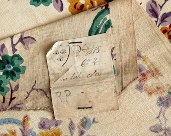 May include: A close-up of a fabric swatch with a floral pattern in shades of yellow, blue, green, and purple. The fabric is beige and has a linen texture. A handwritten tag is attached to the fabric with the text "2m 415 6 = 2  two toile colors RP".