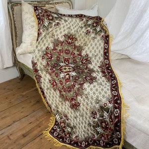 Puede incluir: Una manta de tiro de color crema con un patrón floral rojo, verde y dorado. La manta tiene un borde de flecos y está drapeada sobre una cama blanca.