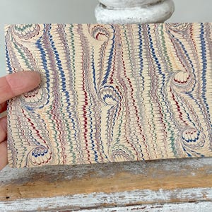 May include: A rectangular piece of paper with a marbled design. The pattern features swirls of red, blue, green, and yellow on a cream-colored background. The paper is held by a hand, showcasing the intricate details of the artwork.