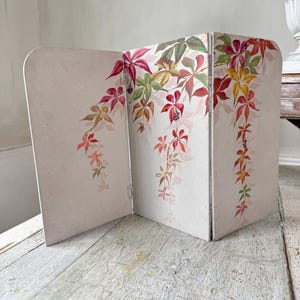 May include: A three-panel folding screen with a cream background. It features a watercolour illustration of autumn leaves in red, green, and yellow. The leaves are depicted cascading down the panels, creating a decorative effect.