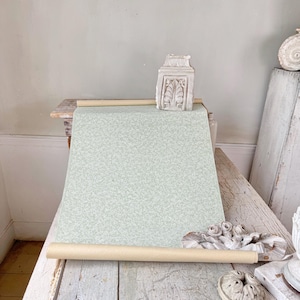 Op de afbeelding: Een rol lichtgroen behang met een subtiel bloemmotief ligt op een verweerde houten tafel. Het behang is opgerold rond een beige kartonnen koker. Decoratieve architecturale elementen zijn rond het behang geplaatst.