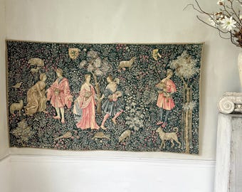 May include: A large tapestry depicting a medieval scene with figures in colorful robes, animals, and foliage. The tapestry is predominantly dark green with accents of pink, red, and gold. It hangs on a light-colored wall.