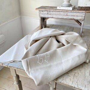 May include: A white linen tablecloth with embroidered initials "GP" in a simple, elegant font. The tablecloth is folded and draped over a rustic wooden table.