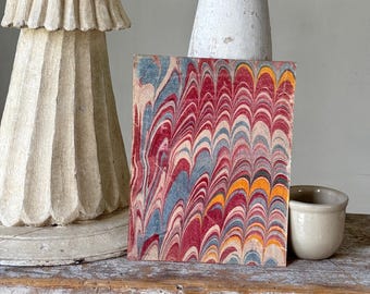 Può includere: Un pezzo rettangolare di carta marmorizzata con un vivace motivo di vortici rossi, blu, gialli e bianchi. La carta è esposta su una superficie di legno invecchiato, accanto a una piccola tazza di ceramica bianco sporco e oggetti decorativi.
