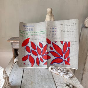 Puede incluir: Un libro antiguo abierto con un diseño floral rojo y azul. El libro tiene páginas de papel cuadriculado con notas manuscritas y el número 5065. El diseño floral está compuesto por formas de hojas rojas delineadas en azul.