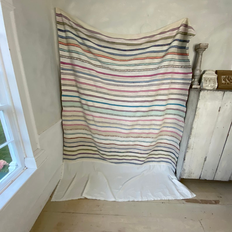 May include: A white striped throw blanket with a variety of colors, including pink, blue, green, and brown. The blanket is hanging on a white wall and has a white fringe around the bottom.