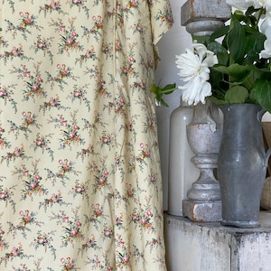 May include: A yellow fabric with a floral pattern of pink and blue flowers. The fabric is draped over a white surface and a wooden stool. A vase with white flowers is in the background.