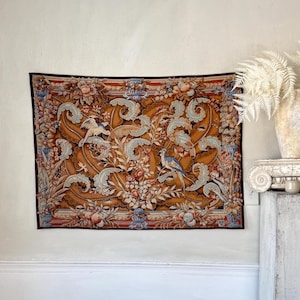 May include: A rectangular tapestry with a brown background, featuring a detailed floral and bird design. The tapestry has a dark border and includes elements of blue, white, and peach. The design includes two birds and various floral arrangements.