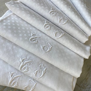 May include: A stack of white linen napkins with embroidered initials. The napkins have a subtle polka dot pattern.