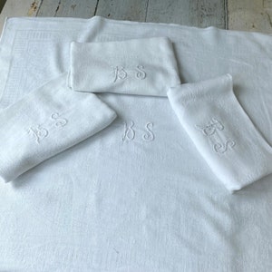 May include: Four white towels with embroidered initials "BS" in a cursive font. The towels are arranged on a white surface.