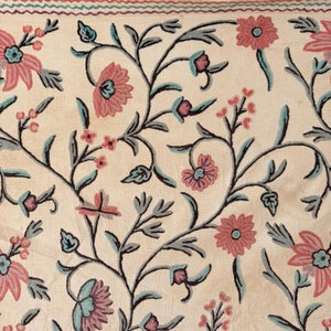 May include: A cream-colored fabric with a floral embroidery pattern. The flowers are pink and blue with a detailed design. The fabric has a subtle sheen.