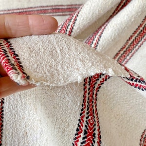 May include: White woven fabric with red and black stripes. The fabric has a textured surface and is folded in half.