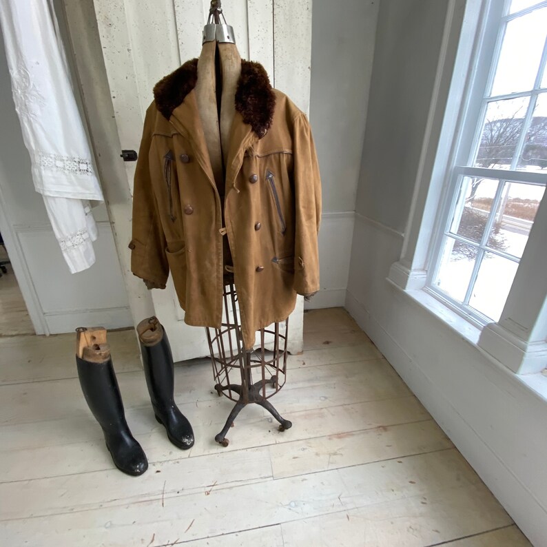 May include: A brown leather jacket with a fur collar on a vintage metal mannequin stand. Two black leather riding boots are on the floor.