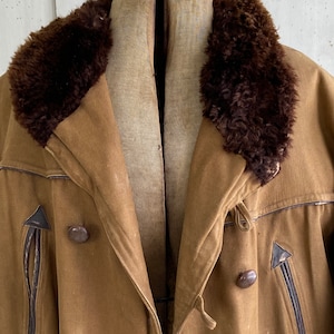 May include: A brown, vintage-style jacket with a fur collar. The jacket has a double-breasted closure with two buttons and a leather trim around the edges.