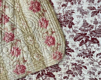 May include: A quilted fabric with a floral pattern in shades of pink and cream. The fabric is folded over itself, revealing a second layer of fabric with a floral pattern in shades of burgundy and white.