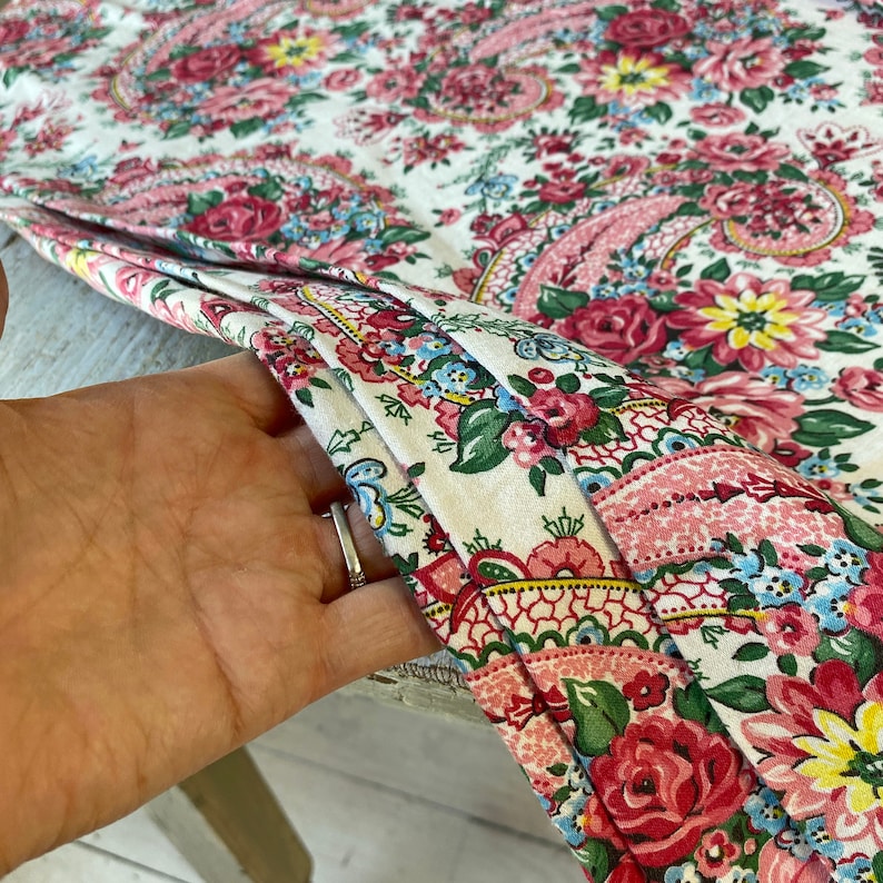 May include: Close-up of a floral fabric with a paisley design. The fabric features a white background with a pattern of red roses, pink flowers, and blue accents. The fabric is folded, revealing the detailed print.