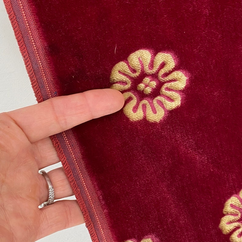 以下が含まれることがあります： 刺繍された金色の花の模様が施された、バーガンディ色のベルベット生地のクローズアップ。生地の端には、バーガンディと金の細いボーダーが付いた装飾的なトリムがあります。指が刺繍された花の1つを指しています。