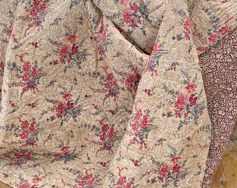 Puede incluir: Una manta de tiro acolchada beige con un patrón floral en tonos de rosa, morado y verde. La manta está hecha de una tela suave y ligera y tiene un aspecto vintage.