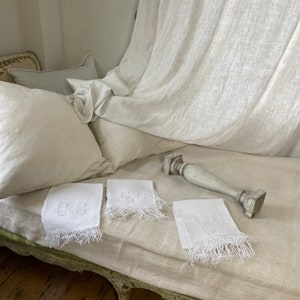 May include: Three white linen hand towels with embroidered initials and fringed edges are displayed on a light beige linen bedspread. A white wooden decorative piece is also on the bedspread.