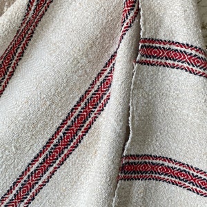 May include: A white linen fabric with red and black woven stripes. The fabric has a rough texture and is slightly frayed at the edges.