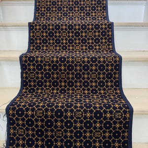 Op de afbeelding: Marineblau traploper met een herhalend gouden geometrisch patroon. De loper is geïnstalleerd op een witte trap. Het patroon bevat cirkels, sterren en de gestileerde letter "M".