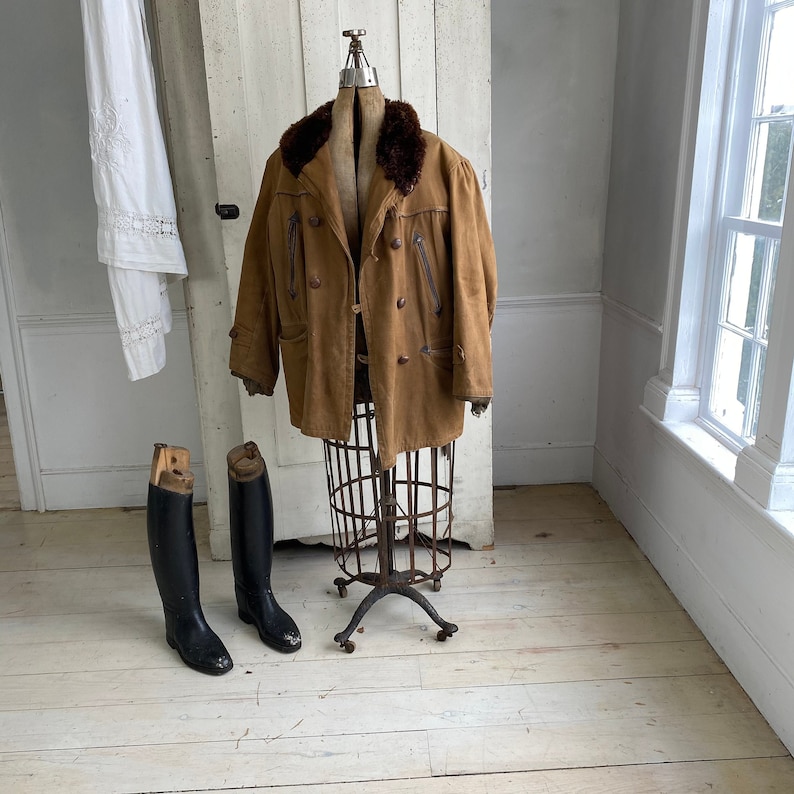 May include: A brown leather jacket with a fur collar on a vintage clothing display stand. Two black leather riding boots are on the floor in front of the stand.