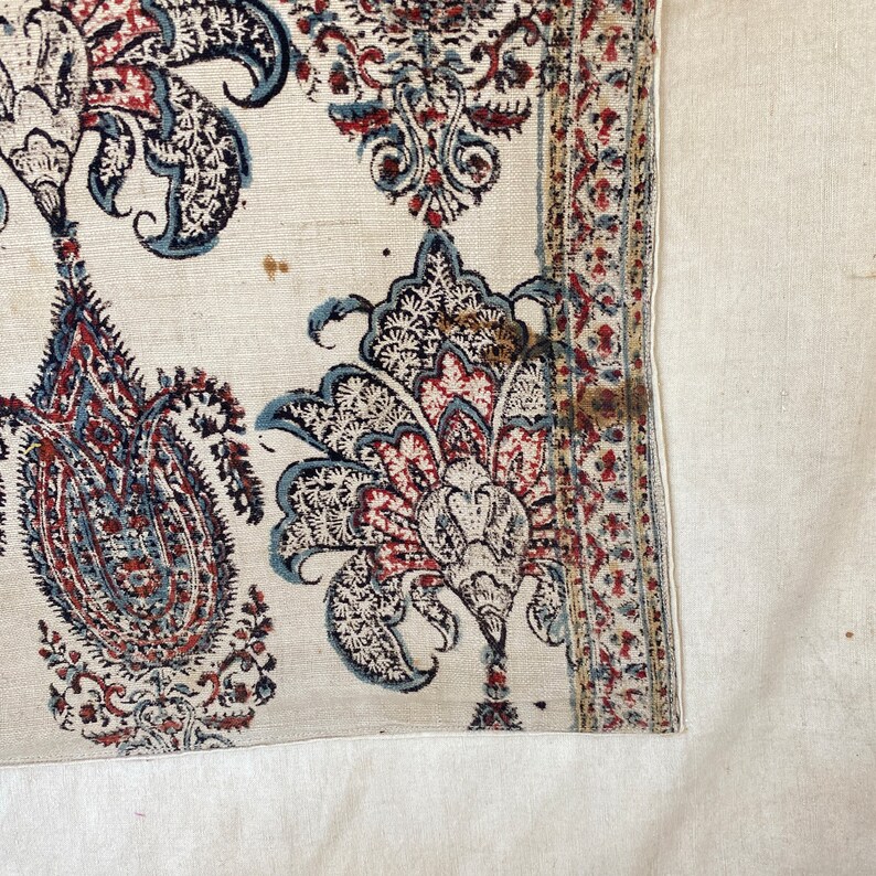 Pu&ograve; includere: Primo piano di un tessuto bianco con un motivo paisley in tonalit&agrave; di blu, rosso e marrone. Il tessuto ha un bordo con un motivo ripetitivo.