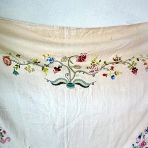May include: A white linen tablecloth with embroidered floral designs in shades of pink, blue, green, and yellow. The embroidery features flowers, leaves, and butterflies.