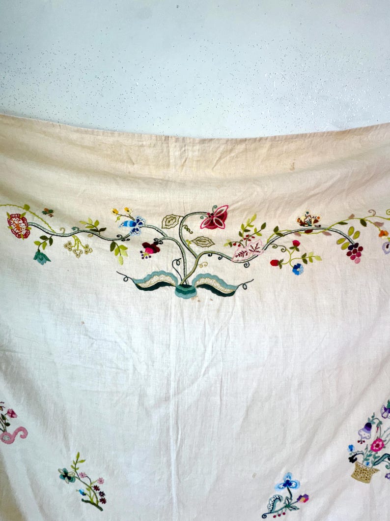 May include: A white linen tablecloth with embroidered floral designs in shades of pink, blue, green, and yellow. The embroidery features flowers, leaves, and butterflies.